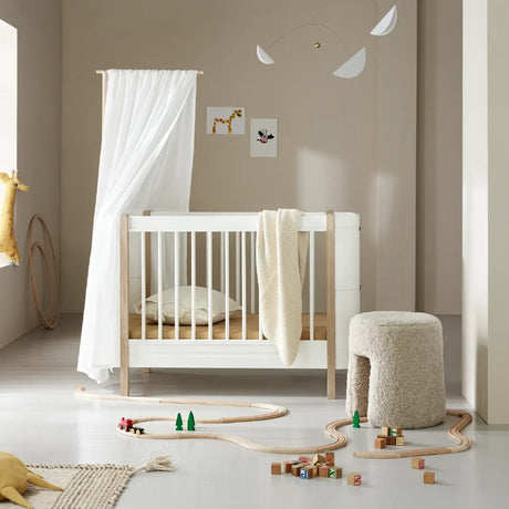 Minimalist nursery setup featuring the Oliver Furniture cot in white and natural wood oak tones, styled with a cosy cream throw, neutral bedding, and a delicate canopy. The room is accented with a fluffy textured stool, wooden train tracks, and colorful blocks, creating a serene and playful ambiance.